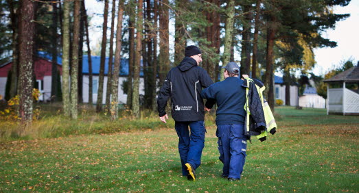 Två personer i arbetskläder som går med ryggen åt kameran på en gräsmatta. Den ena personen håller den andra under armen