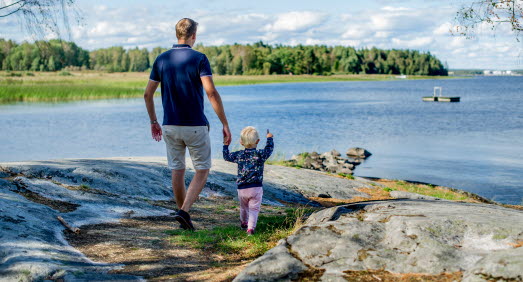Pappa och barn håller varandra i handen och blickar ut över vattnet. Barnet pekar ut mot vattnet. 